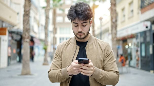 Молодой латиноамериканец использует смартфон с серьезным выражением лица на улице - Фото, изображение