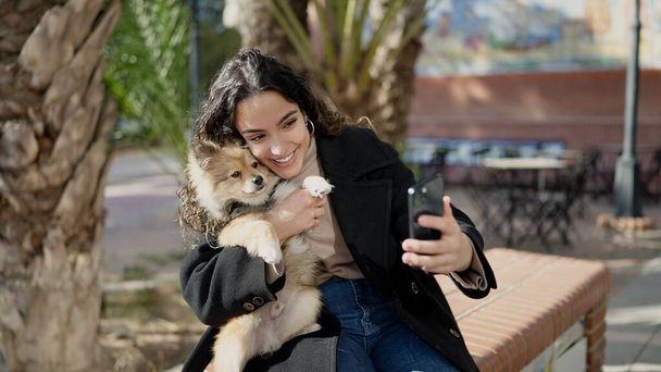 Mladá hispánská žena se psem usměvavý sebevědomí, aby selfie smartphone v parku - Fotografie, Obrázek