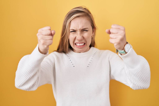 Young caucasian woman wearing white sweater over yellow background angry and mad raising fists frustrated and furious while shouting with anger. rage and aggressive concept.  - Foto, Imagem