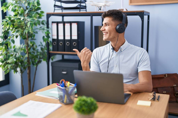 Jeune homme hispanique travaillant au bureau portant des écouteurs souriant avec le visage heureux regardant et pointant vers le côté avec le pouce vers le haut.  - Photo, image