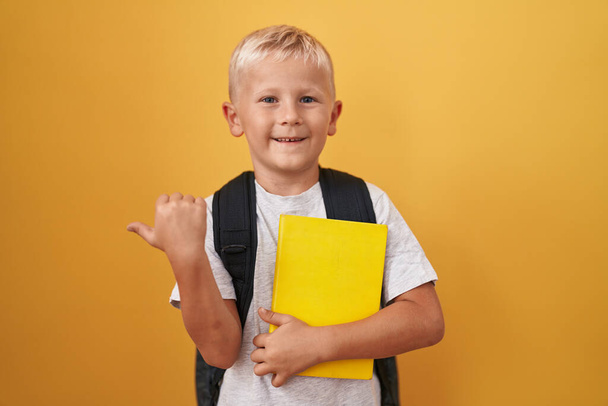 Маленький білий хлопчик в студентському рюкзаку і тримає книгу вказуючи великий палець в сторону усміхнений щасливий з відкритим ротом
  - Фото, зображення