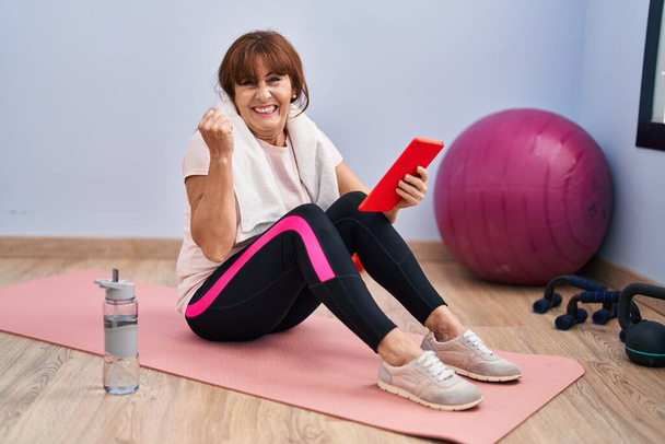 Mujer hispana de mediana edad haciendo ejercicio usando tabletas gritando orgullosa, celebrando la victoria y el éxito muy emocionada con el brazo levantado  - Foto, imagen