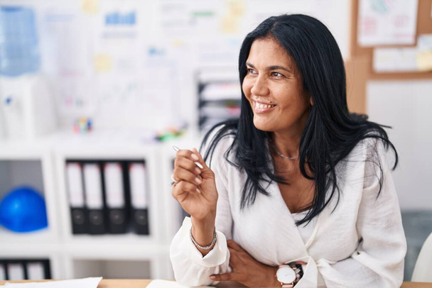 Латиноамериканка среднего возраста, деловая женщина, уверенно улыбающаяся, сидя за столом в офисе - Фото, изображение