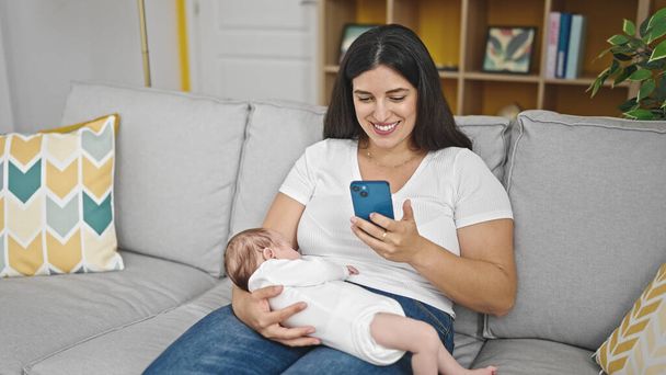 自宅でスマートフォンを使用して赤ちゃんを抱えているソファーに座っている母と赤ちゃんの娘 - 写真・画像