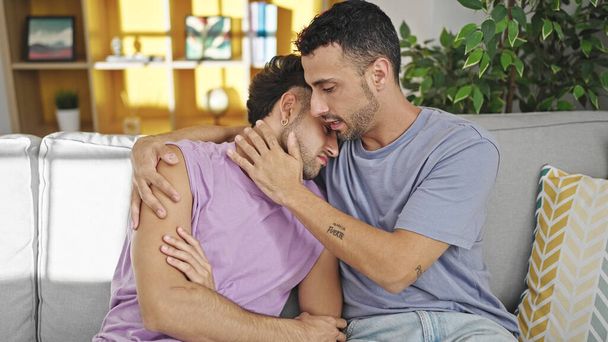 Двое мужчин обнимаются, выглядят расстроенными, поддерживая друг друга дома. - Фото, изображение
