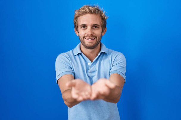 青い背景の上に立っている白人男性は、両手の手のひらで一緒に笑顔を受信またはジェスチャーを与える。保持し保護する  - 写真・画像