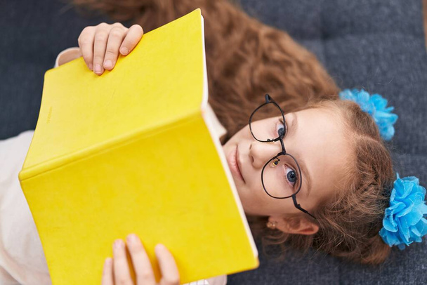 Αξιολάτρευτο καυκάσιος κορίτσι ανάγνωση βιβλίο που βρίσκεται στον καναπέ στο σπίτι - Φωτογραφία, εικόνα