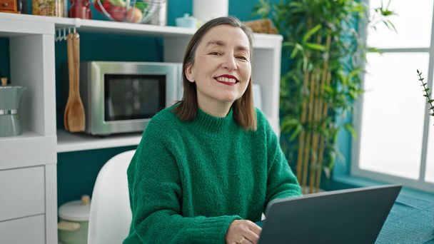 volwassen latino vrouw met grijs haar met behulp van laptop glimlachen in eetkamer - Foto, afbeelding