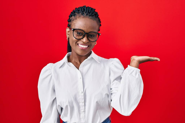 Afrikanerin mit Zöpfen, die vor rotem Hintergrund steht und fröhlich lächelt und mit der Handfläche in die Kamera zeigt.  - Foto, Bild