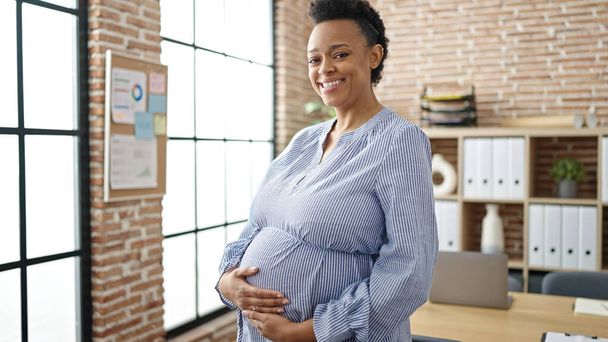 Nuori raskaana oleva nainen liiketoiminnan työntekijä hymyilee luottavainen koskettaa vatsa toimistossa - Valokuva, kuva