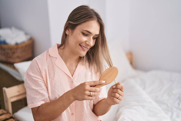 Young caucasian woman combing hair sitting on bed at bedroom - Photo, Image