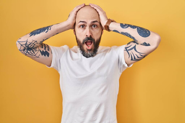 Молодой латиноамериканец с татуировками, стоящий на желтом фоне сумасшедший и напуганный руками на голове, напуганный и удивленный шоком с открытым ртом  - Фото, изображение