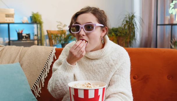 Excited young woman sitting on sofa eating popcorn and watching interesting tv serial, sport game, film, online social media movie content at home. Girl in 3D glasses enjoying domestic entertainment - Photo, Image