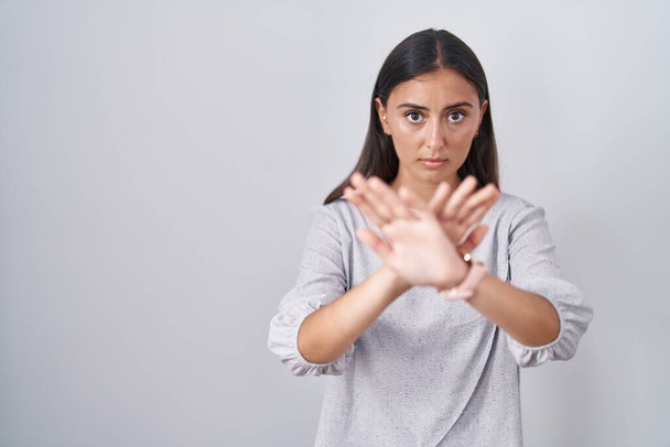Młoda Latynoska kobieta stojąca nad białym tłem odrzucenie wyrażenie skrzyżowanie ramion i dłoni robi negatywny znak, zły twarz  - Zdjęcie, obraz