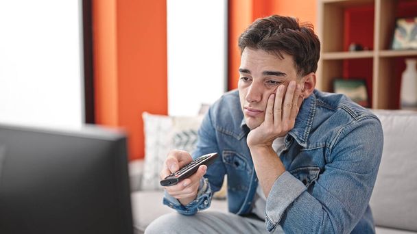 Joven hombre hispano viendo la televisión sentado en el sofá con expresión aburrida en casa - Foto, imagen