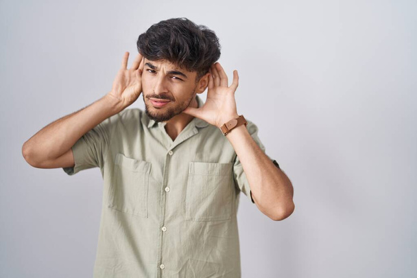 Hombre árabe con barba de pie sobre fondo blanco tratando de escuchar ambas manos en el gesto de la oreja, curioso por chismes. problema auditivo, sordo  - Foto, imagen