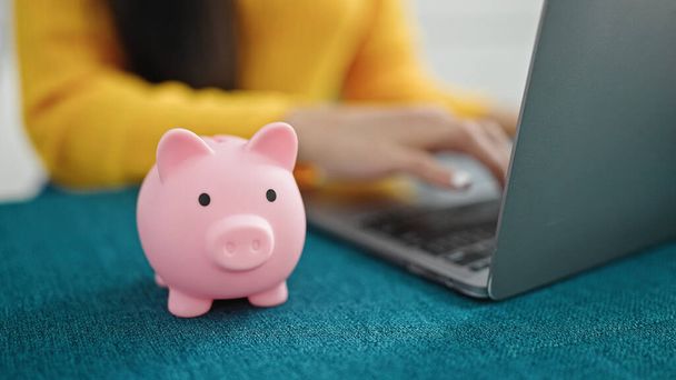 Young beautiful hispanic woman using laptop with piggy bank on table at home - Photo, image
