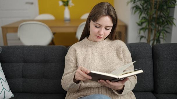 Young blonde woman reading book sitting on sofa at home - 写真・画像