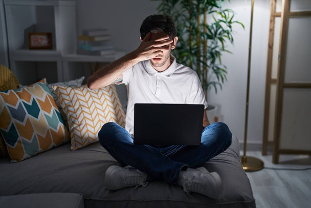 Młody Hiszpan korzystający z laptopa w domu w nocy podglądający w szoku twarz i oczy dłonią, patrzący przez palce ze wstydliwym wyrazem twarzy.  - Zdjęcie, obraz
