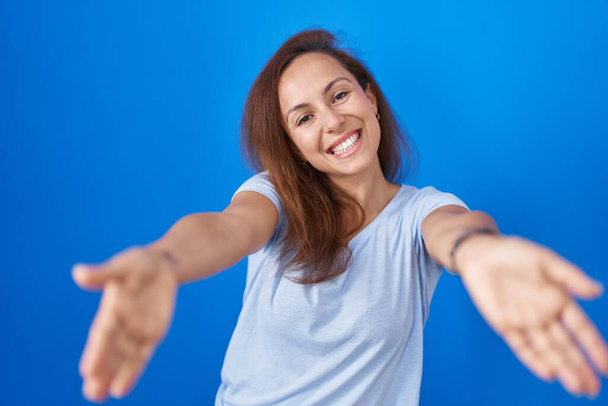 Brunette woman standing over blue background looking at the camera smiling with open arms for hug. cheerful expression embracing happiness.  - Foto, imagen