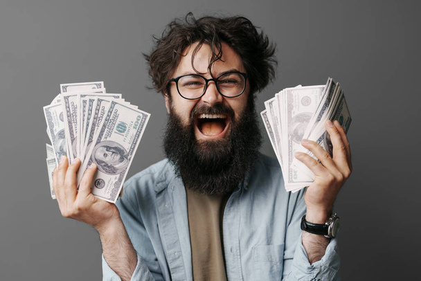 Όμορφος γενειοφόρος άντρας κρατάει τα χρήματα στο χέρι. Ικανοποιημένος γενειοφόρος με τζιν πουκάμισο που δείχνει ένα μάτσο λεφτά απομονωμένα στον γκρίζο τοίχο.. - Φωτογραφία, εικόνα