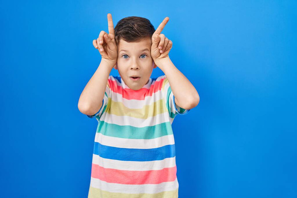 Giovane ragazzo caucasico in piedi su sfondo blu facendo gesto divertente con dito sopra la testa come corna di toro  - Foto, immagini