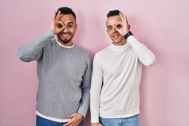Homosexual couple standing over pink background doing ok gesture with hand smiling, eye looking through fingers with happy face.  - Photo, Image