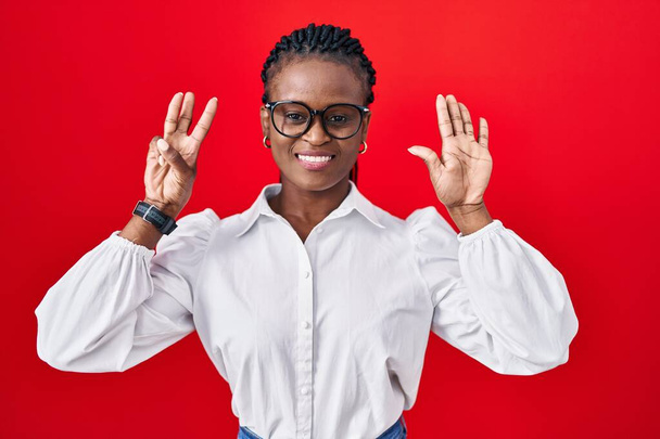 Saçları kırmızı arka planda dikilen Afrikalı kadın sekiz numaralı parmaklarıyla gülümseyerek kendinden emin ve mutlu bir şekilde işaret ediyor..  - Fotoğraf, Görsel