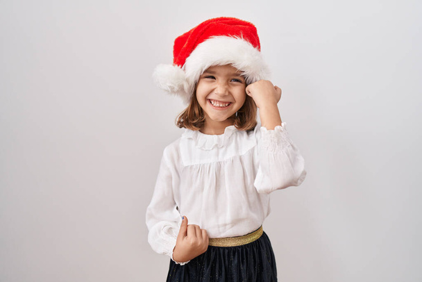 Mała Latynoska dziewczyna w świątecznym kapeluszu świętuje zaskoczona i zdumiona sukcesem z podniesionymi ramionami i zamkniętymi oczami  - Zdjęcie, obraz