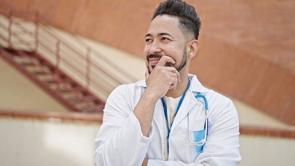 Nuori latino mies lääkäri hymyilee luottavainen ajattelu sairaalassa - Valokuva, kuva