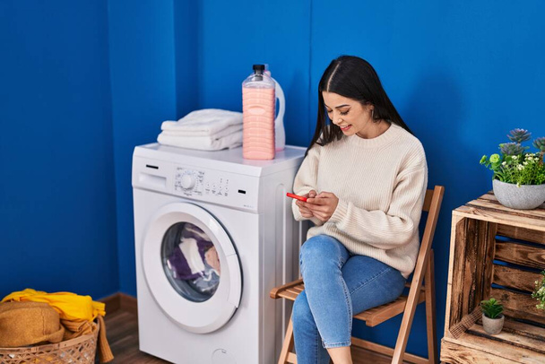 洗濯室で洗濯機を待っているスマートフォンを使用して若い美しいヒスパニック系の女性 - 写真・画像