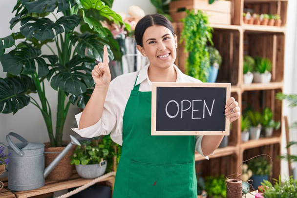 Young hispanic woman working at florist holding open sign smiling with an idea or question pointing finger with happy face, number one  - Photo, Image