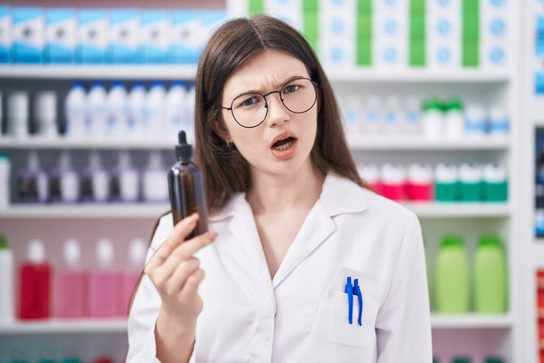 Young caucasian woman working at pharmacy drugstore holding serum scared and amazed with open mouth for surprise, disbelief face  - Photo, Image