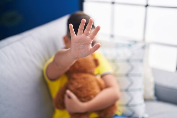 Adorable hispanic boy hugging teddy bear doing stop gesture with hand at home - Photo, Image