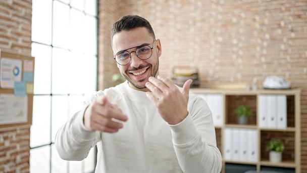 Young hispanic man business worker doing coming gesture at office - Photo, image