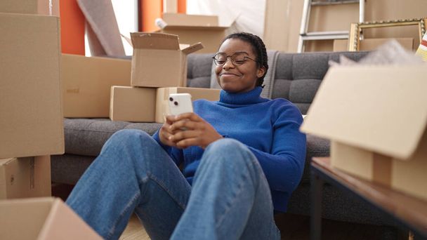 Afrika kökenli Amerikalı kadın akıllı telefon kullanıyor. Yeni evinde yerde oturuyor. - Fotoğraf, Görsel