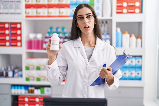Giovane donna bruna che lavora in farmacia con pillole in faccia shock, dall'aspetto scettico e sarcastico, sorpresa a bocca aperta  - Foto, immagini