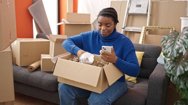 Mujer afroamericana desempacando caja de cartón usando teléfono inteligente en un nuevo hogar - Foto, Imagen