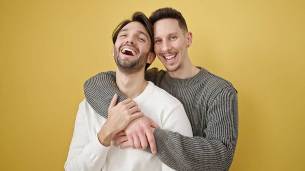 Two men couple hugging each other standing over isolated yellow background - 写真・画像