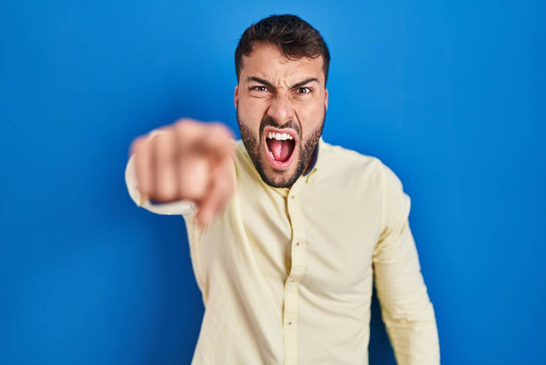Komea latino mies seisoo sininen tausta osoittaa tyytymätön ja turhautunut kameraan, vihainen ja raivoissaan sinulle  - Valokuva, kuva