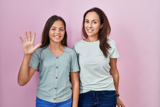 Fiatal anya és lánya áll a rózsaszín háttér mutatja, és rámutatva ujjak száma 5 miközben mosolygós magabiztos és boldog.  - Fotó, kép
