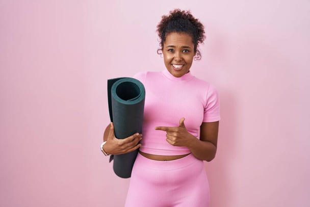 Junge hispanische Frau mit lockigem Haar hält Yogamatte über rosa Hintergrund und zeigt besorgt und nervös mit dem Zeigefinger, besorgt und überrascht  - Foto, Bild