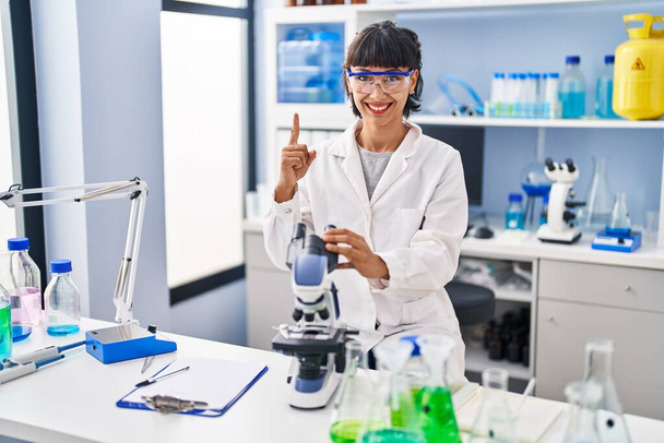 Fiatal spanyol nő dolgozik tudós laboratóriumban mosolyogva egy ötlettel vagy kérdéssel ujjal mutogatva boldog arccal, első számú  - Fotó, kép