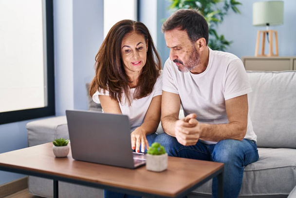 Orta yaşlı erkek ve kadın çift evdeki koltukta dizüstü bilgisayar kullanıyor. - Fotoğraf, Görsel