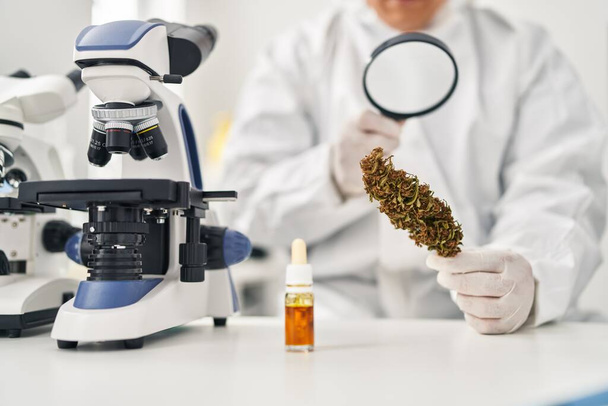 Young latin man scientist looking cannabis herb with loupe at laboratory - Фото, зображення