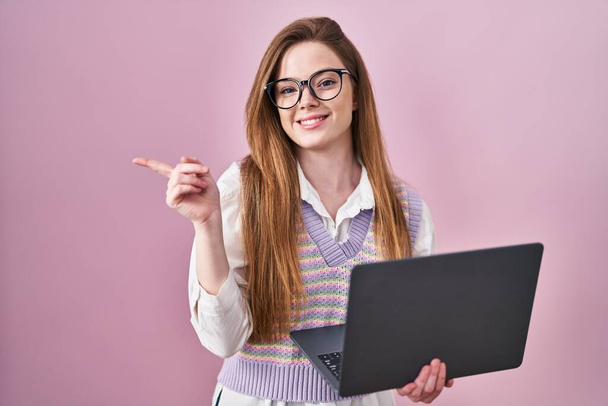 Junge kaukasische Frau, die mit einem Computer-Laptop arbeitet, mit einem breiten Lächeln im Gesicht und zeigt mit dem Finger zur Seite in die Kamera.  - Foto, Bild