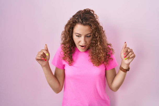 Junge kaukasische Frau, die über rosa Hintergrund steht und nach unten zeigt, mit Fingern, die Werbung zeigen, überraschtem Gesicht und offenem Mund  - Foto, Bild
