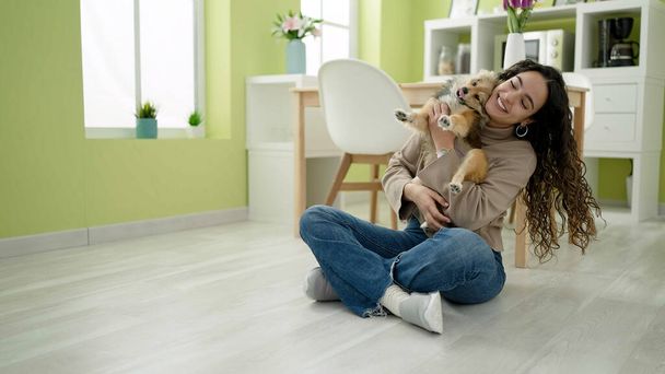 Young hispanic woman with dog sitting on floor hugging at dinning room - Foto, immagini
