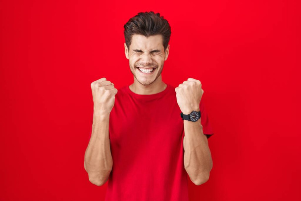 Young hispanic man standing over red background excited for success with arms raised and eyes closed celebrating victory smiling. winner concept.  - Fotografie, Obrázek