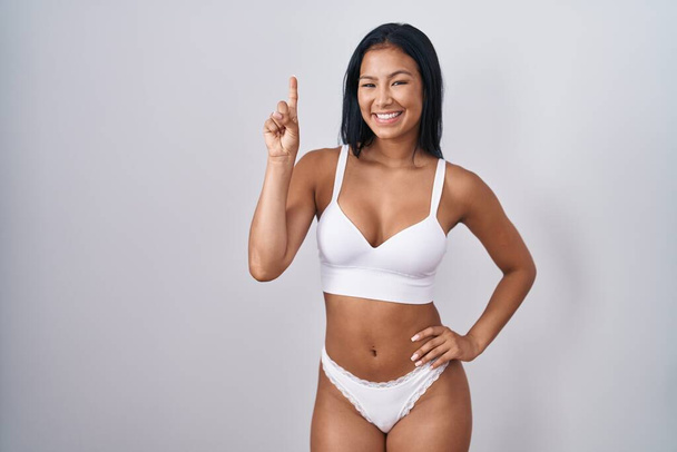 Hispanic woman wearing lingerie showing and pointing up with finger number one while smiling confident and happy.  - Photo, Image